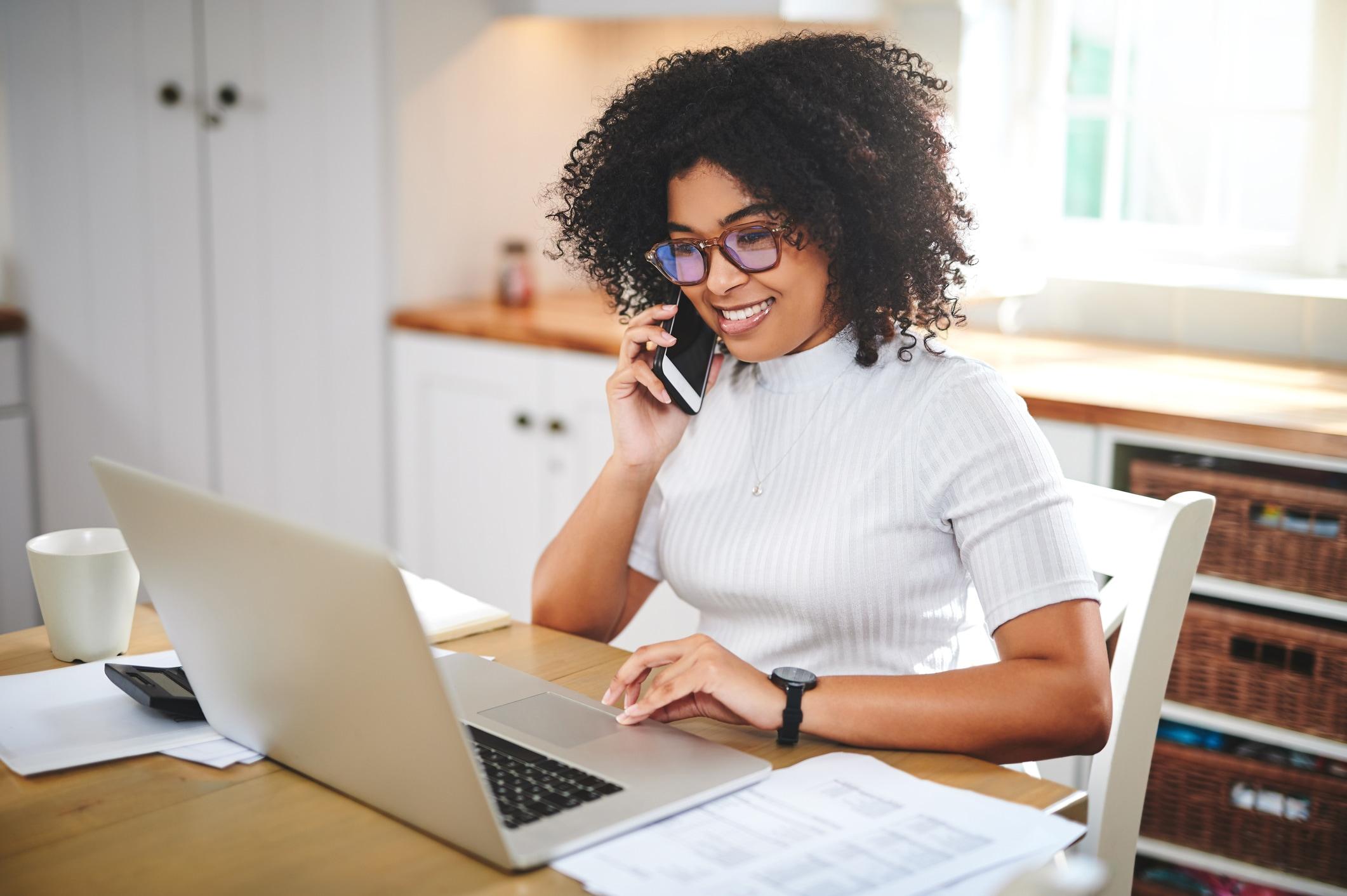 Une femme assise à un comptoir de cuisine, travaillant sur un ordinateur portable et parlant au téléphone.